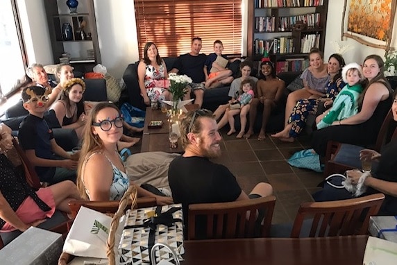 A group of 18 people sit on couches and chairs in a living room, smiling near Christmas presents.