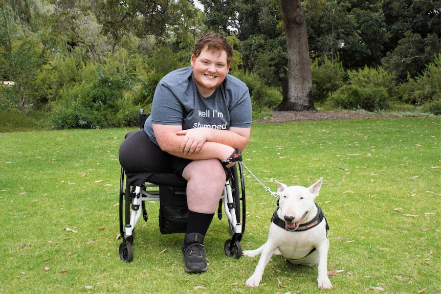 Angus sits in a wheelchair, smiling and holding the lead of Maggie the bull terrier who sits on grass by his side 