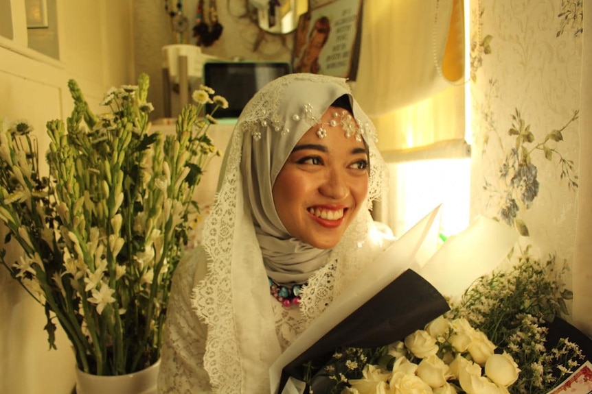 A photo of the bride before her wedding, wearing a headscarf and surrounded by flowers.