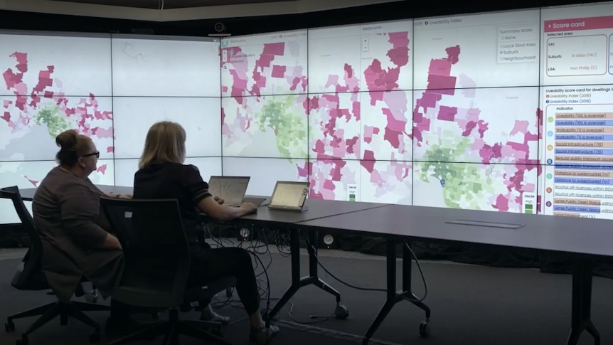 Magda Szubanski and unknown woman sit at laptop computer in front of very large digital wall showing a map