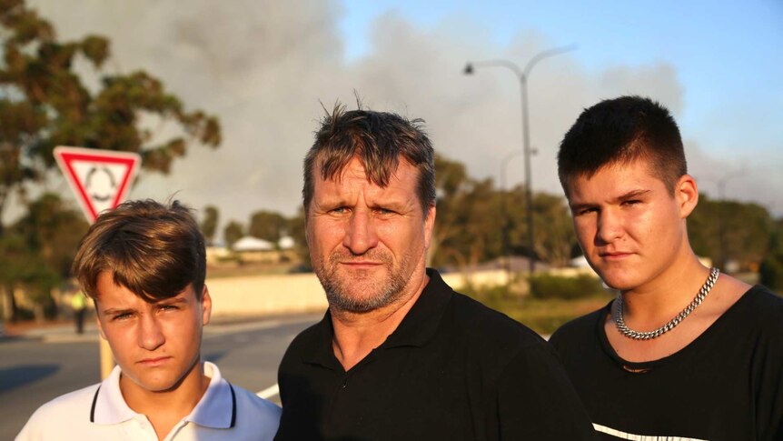 Three men stand on a roadside with flames in background