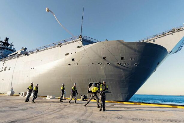 The bow of a large grey navy ship anchored at a wharf.