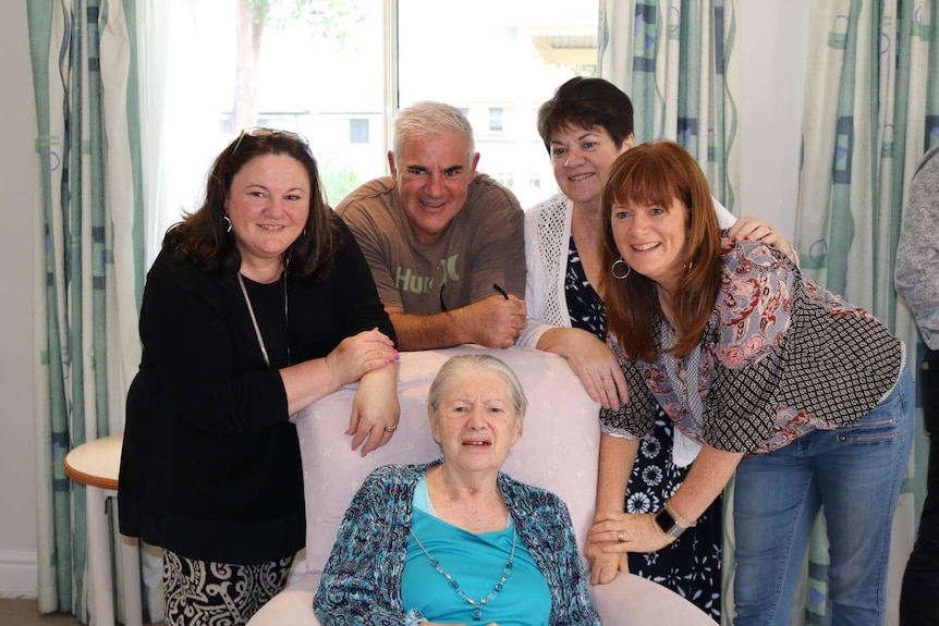 An elderly woman sits in a chair four people surround her, smiling for a photo.