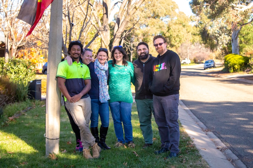 Six adults smile arm in arm next to a flagpole at the top of a street.