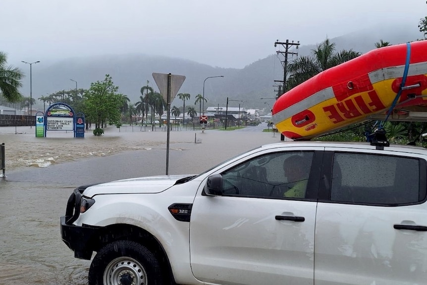 An emergency services ute with a boat on the roof is parked in front of a flooded road.