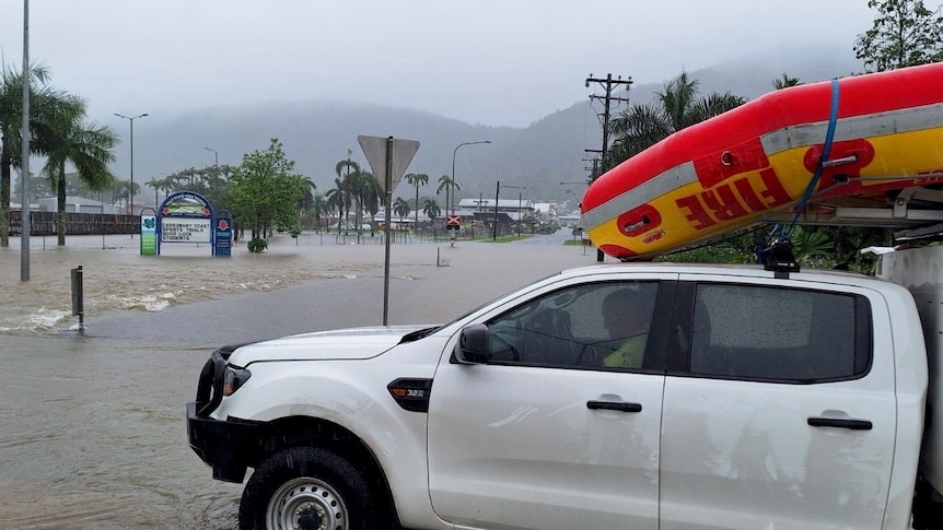 An emergency services ute with a boat on the roof is parked in front of a flooded road.