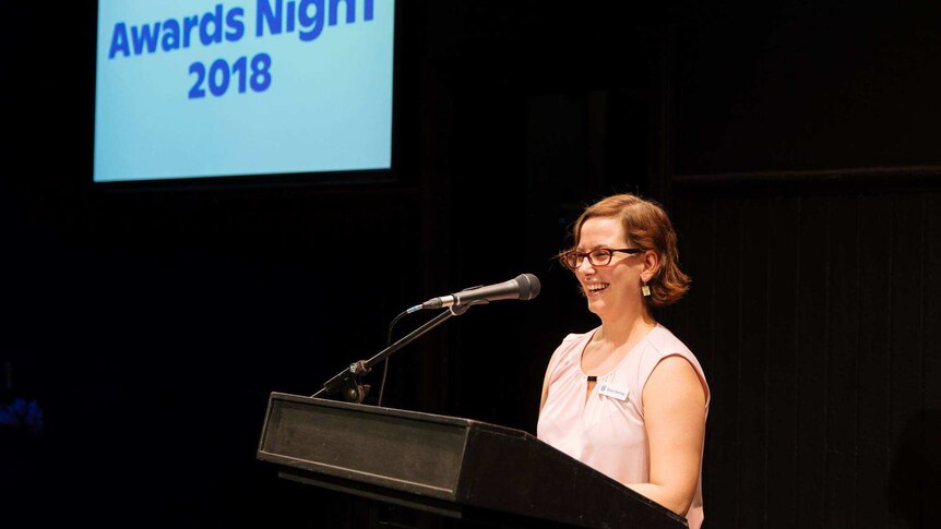 Kathryn Osbourne stands at a podium laughing, a projection says "awards night 2018" behind her.