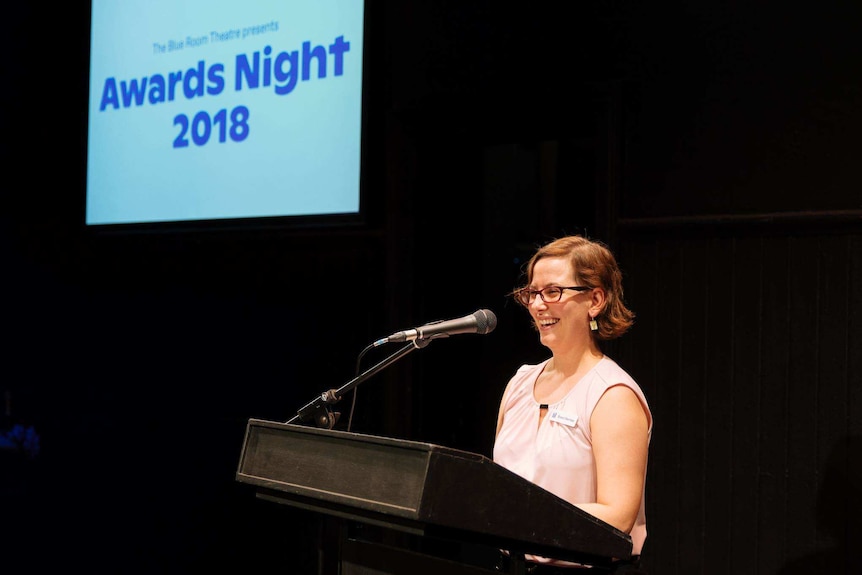 Kathryn Osbourne stands at a podium laughing, a projection says "awards night 2018" behind her.