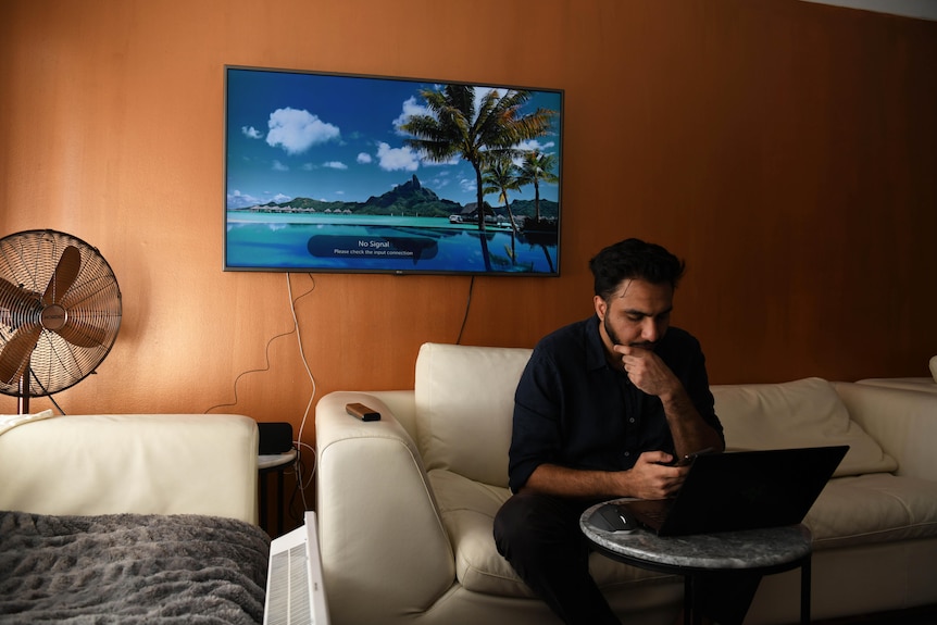 A 26-year-old man with dark hard and trimmed beard sits on a white counch looking down on his mobile phone with laptop on table