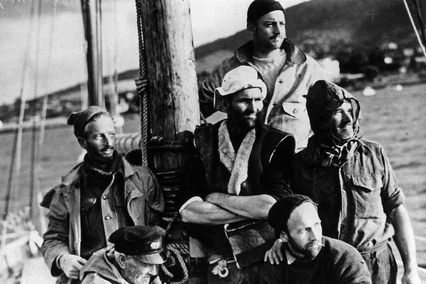 A group of sailors on board a yacht stand or sit looking out to sea.