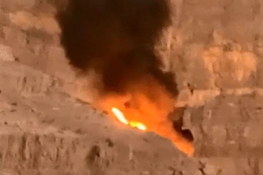 Flames burn and smoke billows from mountain crevice following helicopter crash.
