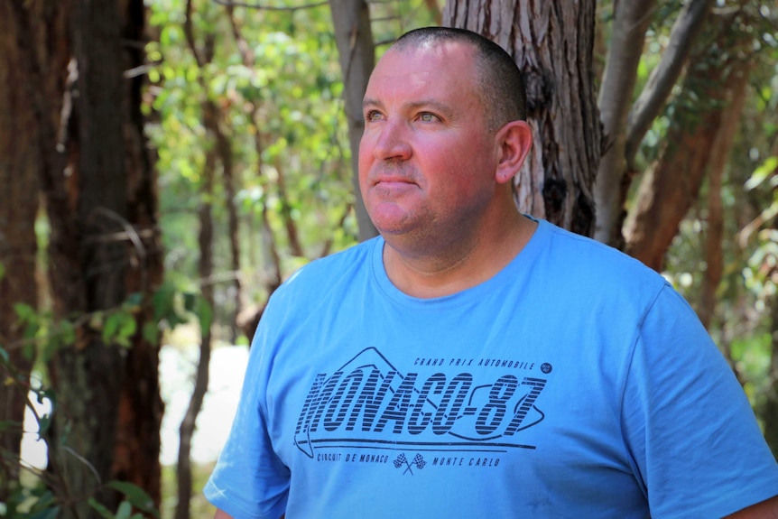 A mid shot of a man wearing a blue t-shirt standing in the bush looking off camera.