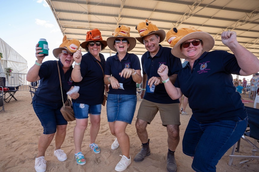 A group of men and women in matching blue t-shirts ear colorful hats, holding drinks pump their fists and pose for a photo.
