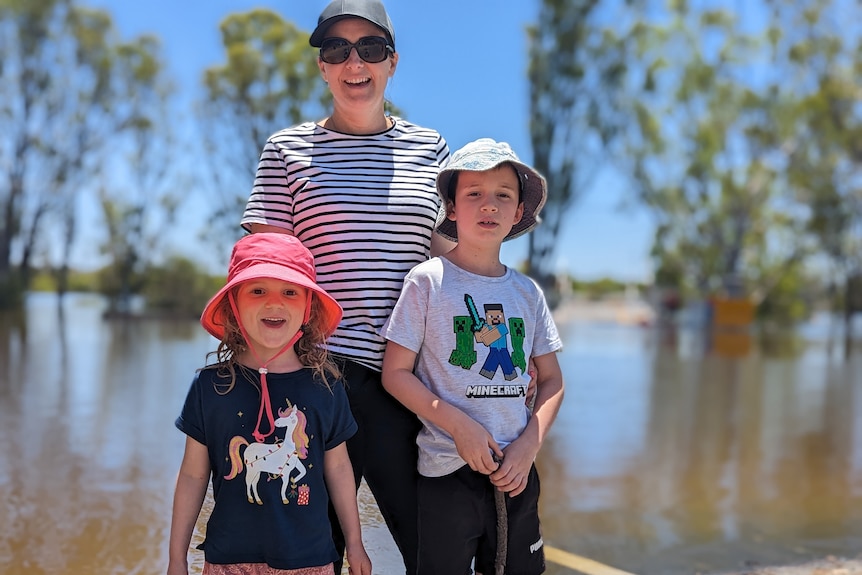 A young white woman stands with two young children smiling next to the floodwaters in Swan Reach.