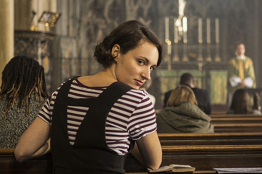 A young woman wearing a striped t-shirt and black tunic over the top looks over her shoulder while kneeling at a church pew.