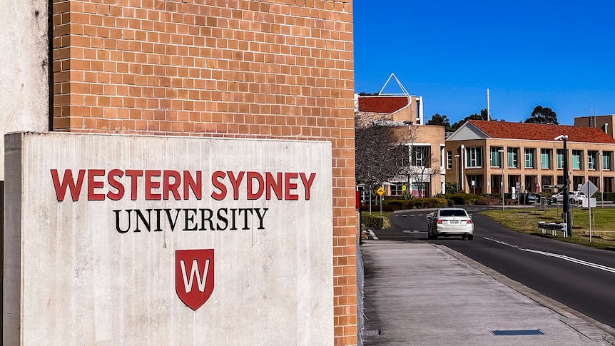 A sign on a brick wall reads 'Western Sydney University', in the distance are some large buildings.