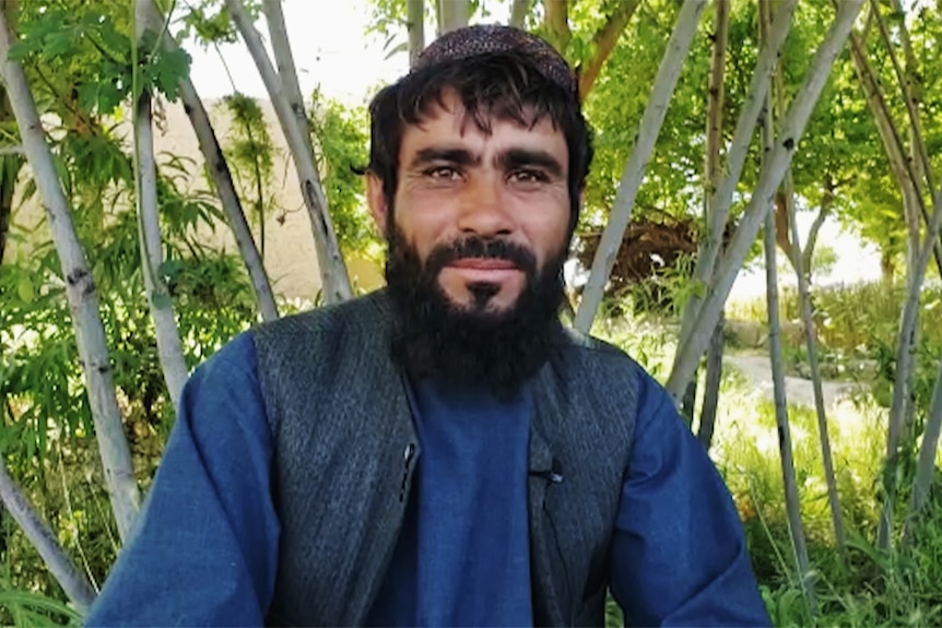 An Afghan man seated in front of green trees.