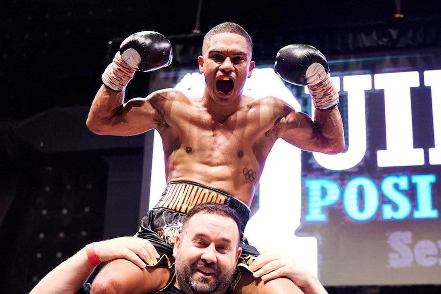 A boxer sitting on the shoulders of another man in a boxing ring.