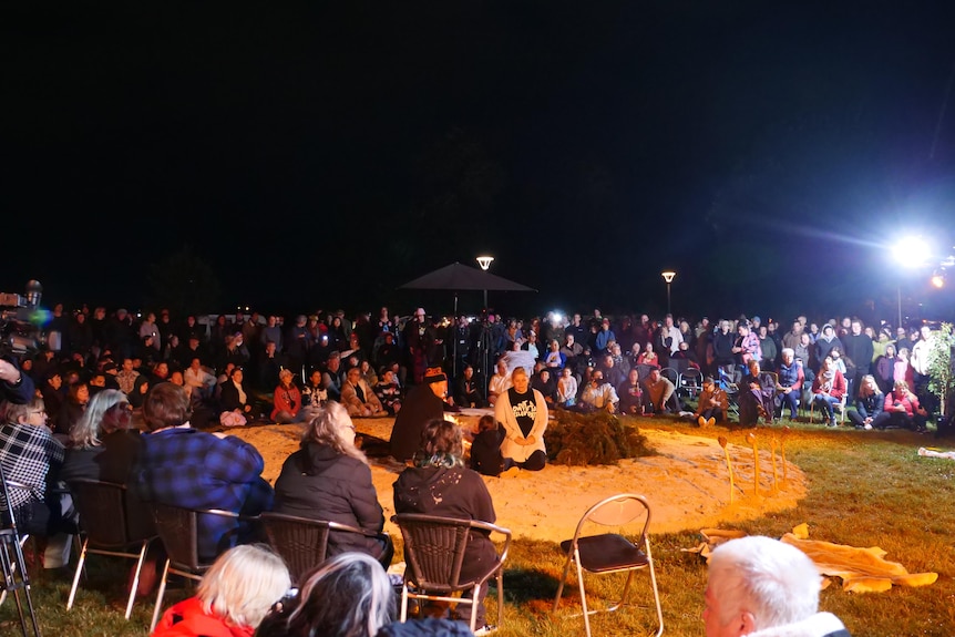 A very large crowd sits on the grass of the park in the pre-dawn darkness.