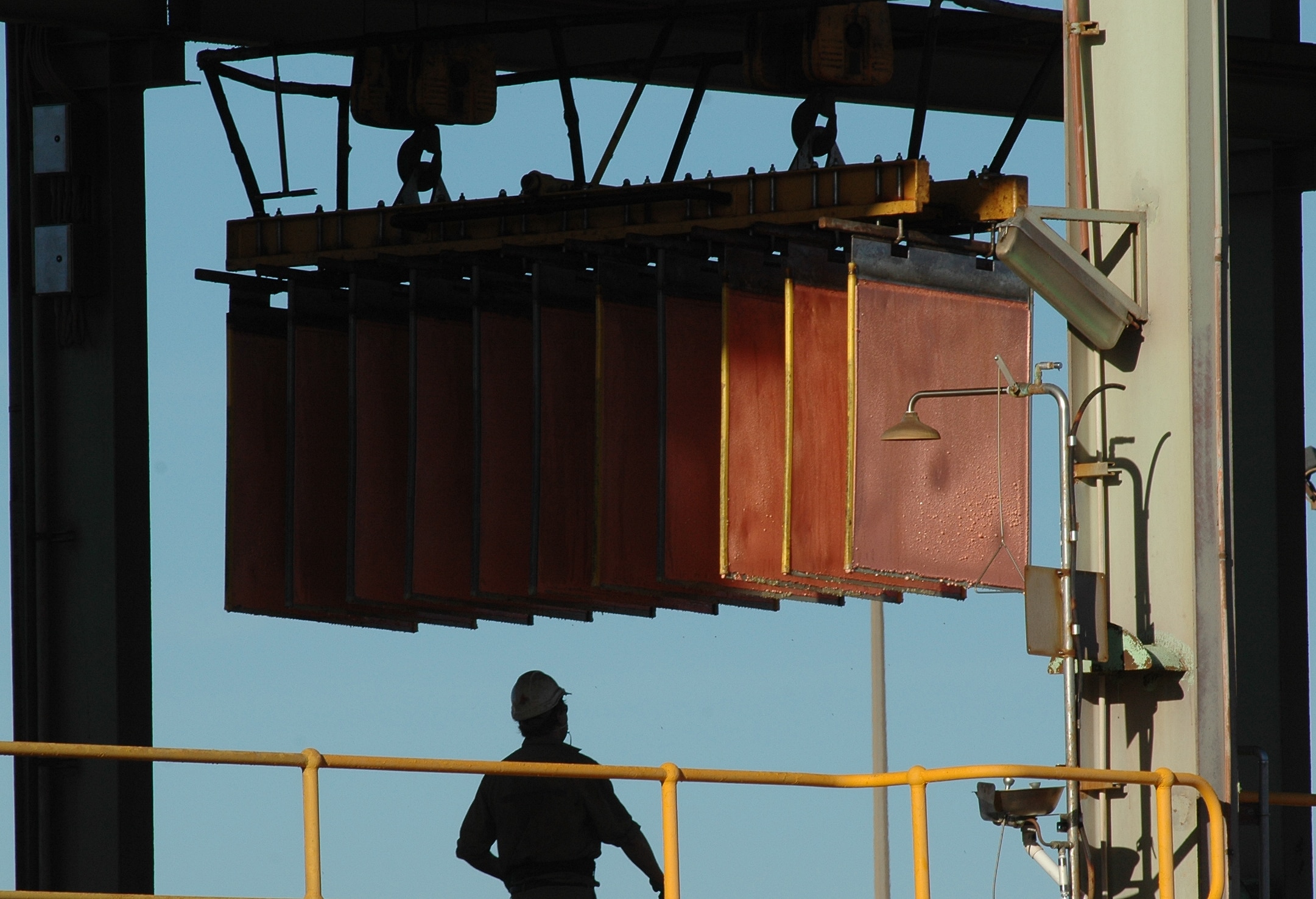 a large claw clasps a row of individual copper cathode squares, with a worker at the bottom of the frame dwarfed by the cathodes