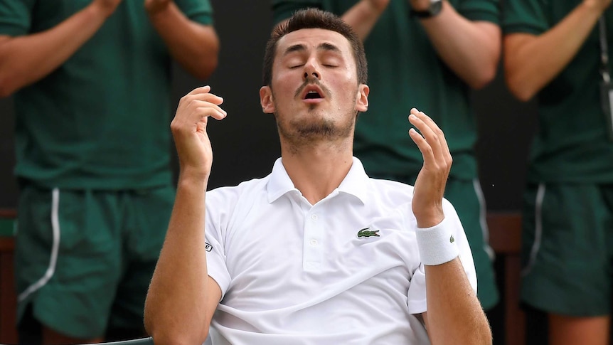 Bernard Tomic raises his hands with his eyes closed while sitting in the player's chair at Wimbledon