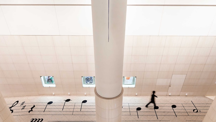 A person walks across the score of Satie's Gymnopédie No. 1 printed on the floor of a large atrium.