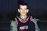 A young man sits on the ground outside at night wearing a hoodie jumper, tracksuit pants, white trainers and an NBA jersey.