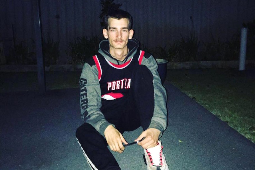 A young man sits on the ground outside at night wearing a hoodie jumper, tracksuit pants, white trainers and an NBA jersey.