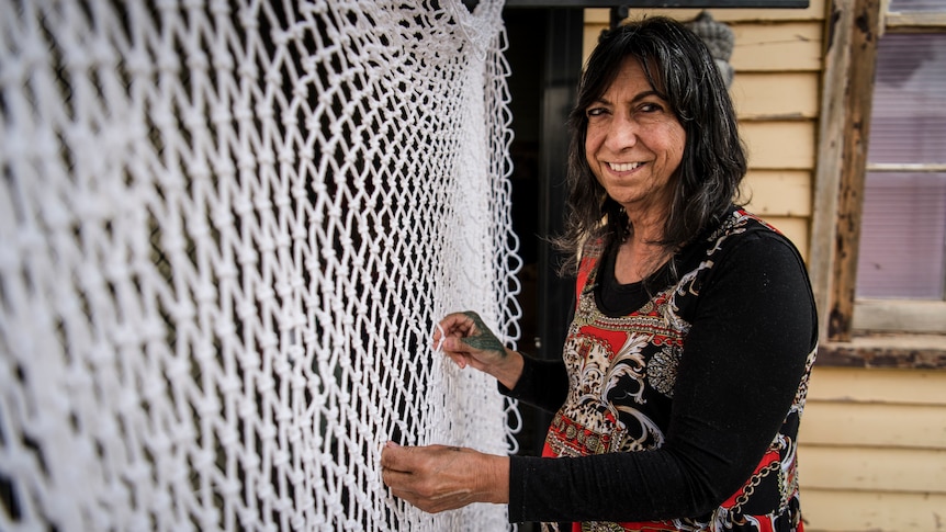 A woman stands with her hands on a white woven artwork that resembles a fishing net.