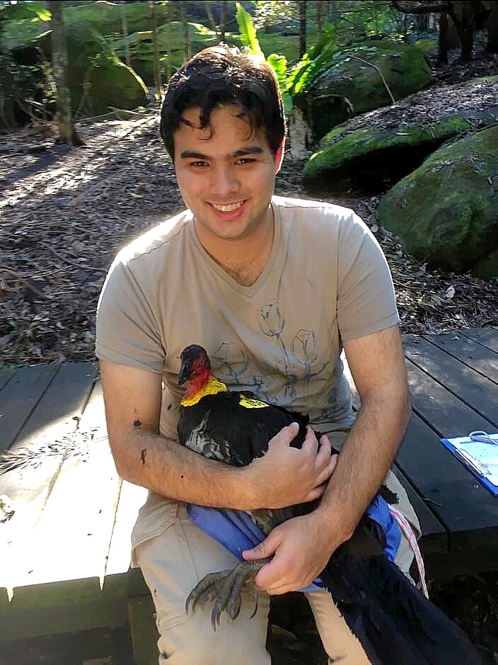 A man in a beige t-shirt sitting down in a park environment holding a black bird with a red featherless head 