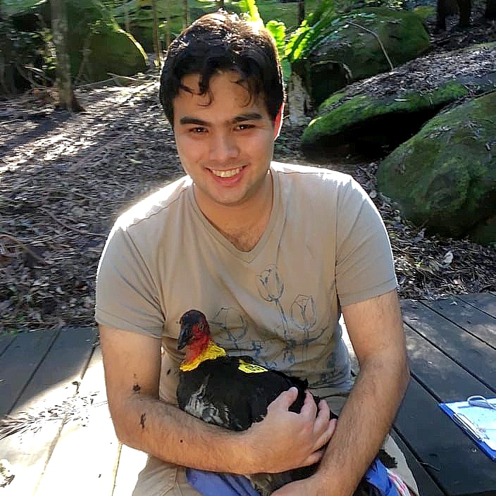 A man in a beige t-shirt sitting down in a park environment holding a black bird with a red featherless head 