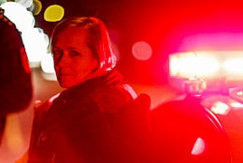 Shot of Sarah standing in front of police car with red light.