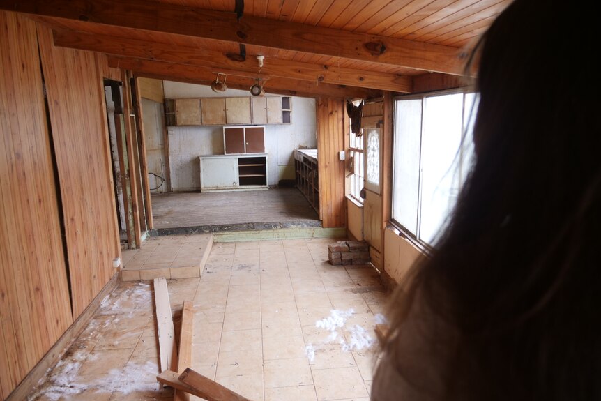 a woman with long hair, gray t-shirt and jeans looks at her newly purchased wooden house that cannot be lived in