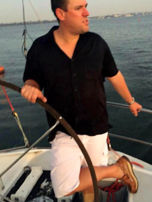 Mr Edman stands at the wheel of a yacht, holding the wheel, in boat shoes, white shorts and a shirt. He looks into the distance.