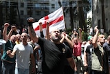 A group of men gathered in a street raise their fists into the air with a Georgian flag in the background.