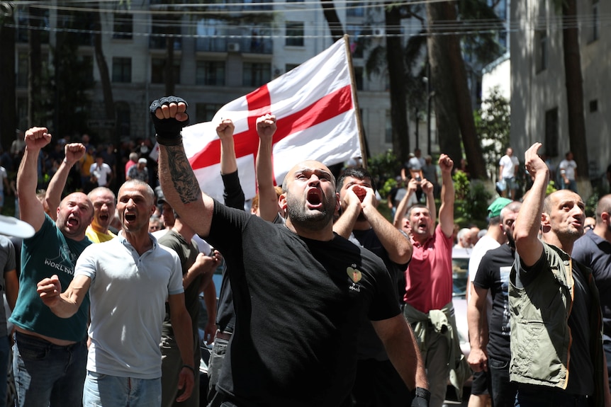 A group of men gathered in a street raise their fists into the air with a Georgian flag in the background.