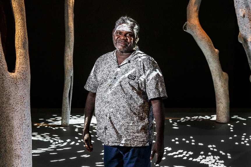 Man standing in darkened gallery surrounded by painted upright hollow logs, white ochre across his forehead.