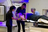 Man lies prone on table of hospital machine with two staff helping him