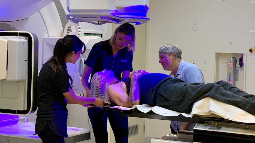 Man lies prone on table of hospital machine with two staff helping him