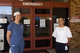 a female doctor and a female nurse manager in uniform standing outside the sliding door entrance to a hospital