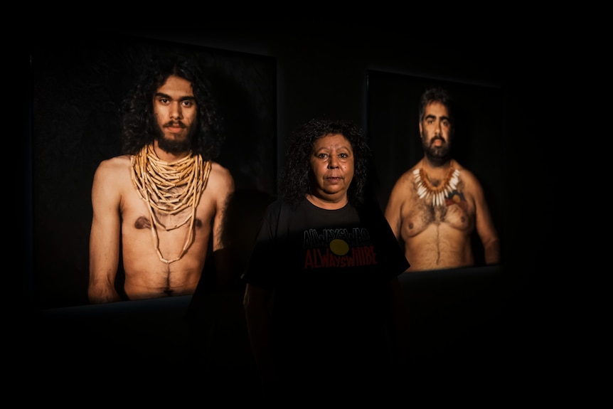 Maree Clarke stands in front of photographs of Aboriginal men, who are posed shirtless wearing traditional necklaces