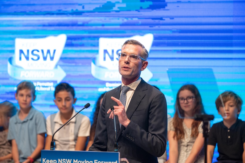 A man wearing glasses standing behind a podium talking with a number of children sitting behind him on a stage.