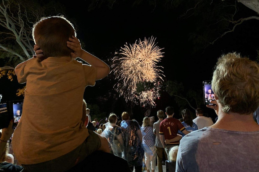 A young boy on his Dad's shoulders covers his ears as he watches fireworks.
