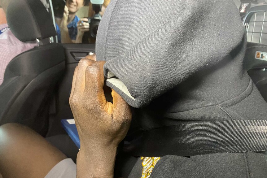 A handcuffed African man covers his face with a hoodie in back of a car with photographers behind.