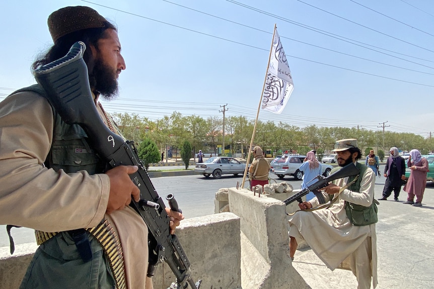 A Taliban fighter stands on top of a guard tower, holding a gun. A white flag is visible behind him