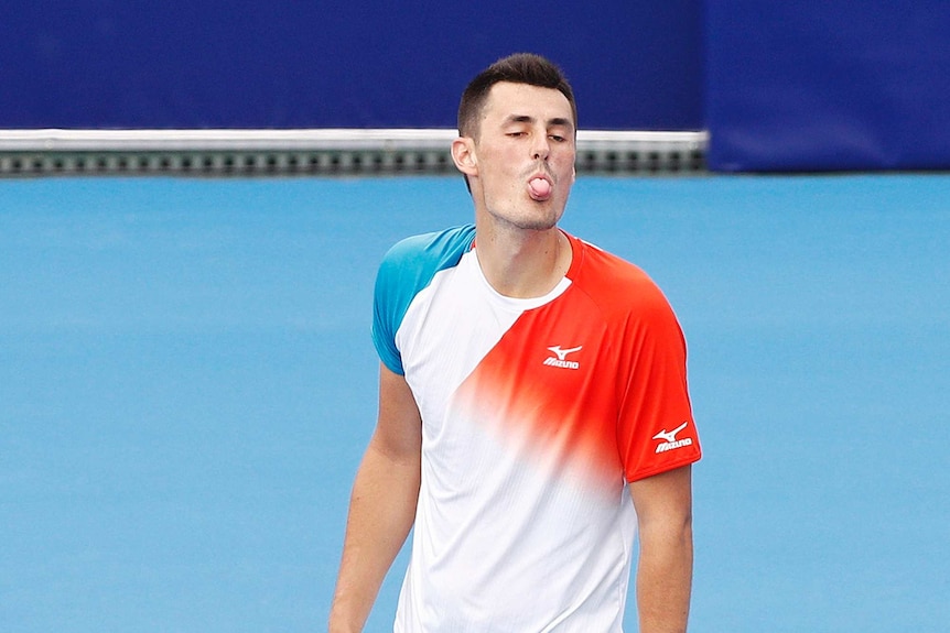 A man wearing a white shirt with red and blue streaks sticks his tongue out while holding a tennis racquet
