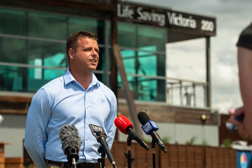 A fit young man in a suit stands in front of lifesaving vic with media mics in front of him