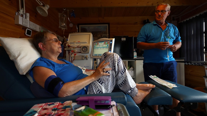 A woman sits in a raised bed undergoing dialysis as her husband speaks to her nearby.