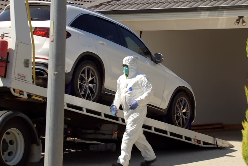 A person in a forensic suit stands next to a white car being towed out of a driveway