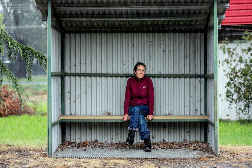 Woman in red jumper and blue jeans with greying black hair sits in tin bush shelter with rain falling